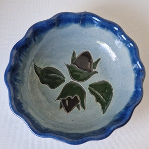 May include: A blue and white ceramic bowl with a scalloped rim. The bowl features a hand-painted design of three stylised flowers with green leaves and black centres.