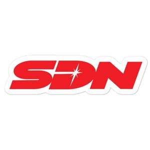 Op de afbeelding: Een gestanste sticker met de letters SDN in een vet, rood, schreefloos lettertype. De letter D bevat in het midden een witte vierpuntige ster. Het gehele ontwerp is voorzien van een dunne witte rand tegen een effen achtergrond.