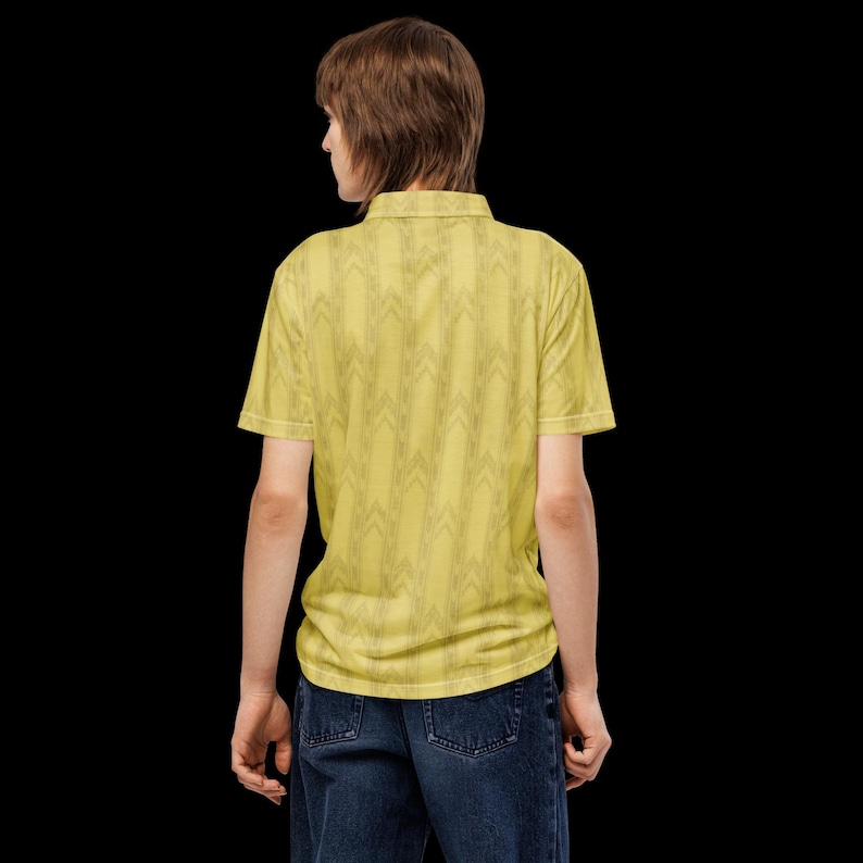 May include: A short-sleeved polo shirt in a pale yellow hue featuring a repeating dark gray chevron pattern throughout the fabric. The shirt has a classic collar and is paired with dark blue denim jeans, visible from the back view against a black background.