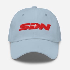 Op de afbeelding: Lichtblauwe baseballcap met de geborduurde rode letters "SDN" en een wit sterrenpatroon. De cap heeft een gebogen klep en een verstelbare band aan de achterkant.