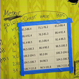 May include: A size chart printed on white paper with blue painter's tape on a yellow background. The chart lists sizes XS through 3XL with corresponding metric measurements for chest, waist, and hips. The text includes 'METRIC', 'CHEST', 'WAIST', 'HIPS', and size labels.