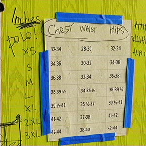May include: A size chart printed on paper and taped with blue painter's tape to a yellow wall. The chart lists sizes XS through 3XL with corresponding chest, waist, and hip measurements in inches. Handwritten text includes "Inches" and "Polo!" along the left side.