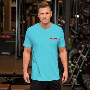May include: A light blue t-shirt with short sleeves. The shirt has the red letters "SDN" printed on the upper chest. The person is standing in a gym.