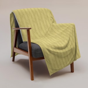 May include: A soft, light yellow throw blanket featuring a repeating chevron pattern in a darker shade of yellow. The blanket is draped over a dark gray armchair with wooden armrests and legs, showcasing its texture and folded fabric drape.