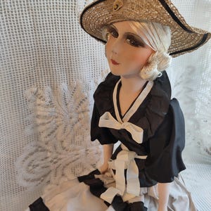 May include: A vintage porcelain doll wearing a straw hat and a black and white dress. The doll has blonde hair and is wearing a wide-brimmed straw hat with a black ribbon. The dress is black with white accents and a white bow.