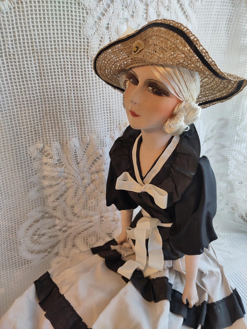 May include: A vintage porcelain doll wearing a straw hat and a black and white dress. The doll has blonde hair and is wearing a wide-brimmed straw hat with a black ribbon. The dress is black with white accents and a white bow.