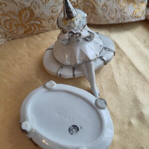 Puede incluir: Caja de porcelana blanca con tapa que presenta una figurita de un buf&oacute;n con un sombrero puntiagudo. La caja tiene un borde plateado y la parte inferior de la caja est&aacute; marcada con el n&uacute;mero "1540".