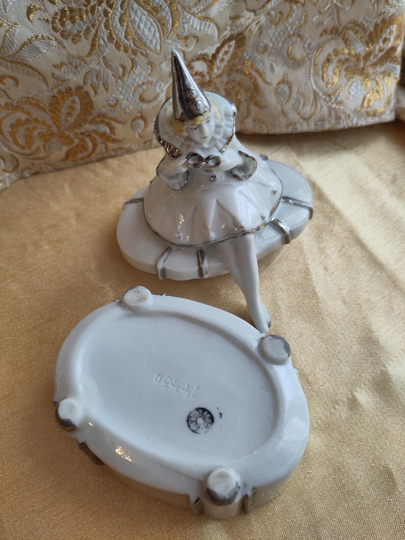 Puede incluir: Caja de porcelana blanca con tapa que presenta una figurita de un buf&oacute;n con un sombrero puntiagudo. La caja tiene un borde plateado y la parte inferior de la caja est&aacute; marcada con el n&uacute;mero "1540".