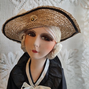 May include: A vintage doll wearing a straw hat with a black ribbon trim and a small flower. The doll has a black and white dress with a bow, and a painted face with detailed features, including red lips and large eyes.