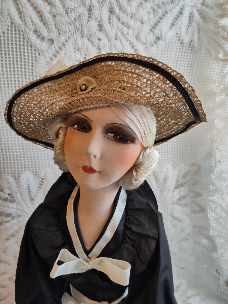 May include: A vintage doll wearing a straw hat with a black ribbon trim and a small flower. The doll has a black and white dress with a bow, and a painted face with detailed features, including red lips and large eyes.