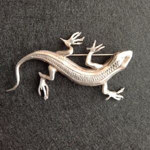 Op de afbeelding: Een zilveren broche in de vorm van een hagedis met een gedetailleerd, getextureerd lichaam en een pin aan de achterkant.