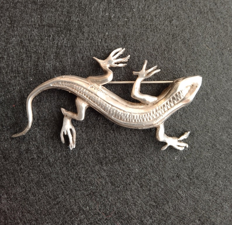Op de afbeelding: Een zilveren broche in de vorm van een hagedis met een gedetailleerd, getextureerd lichaam en een pin aan de achterkant.