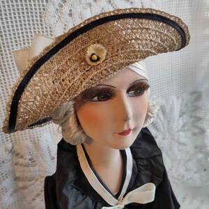 May include: A vintage straw hat with a black ribbon trim and a small floral accent. The hat is part of a doll wearing a black dress with a white bow. The doll has a painted face with red lips and long eyelashes.