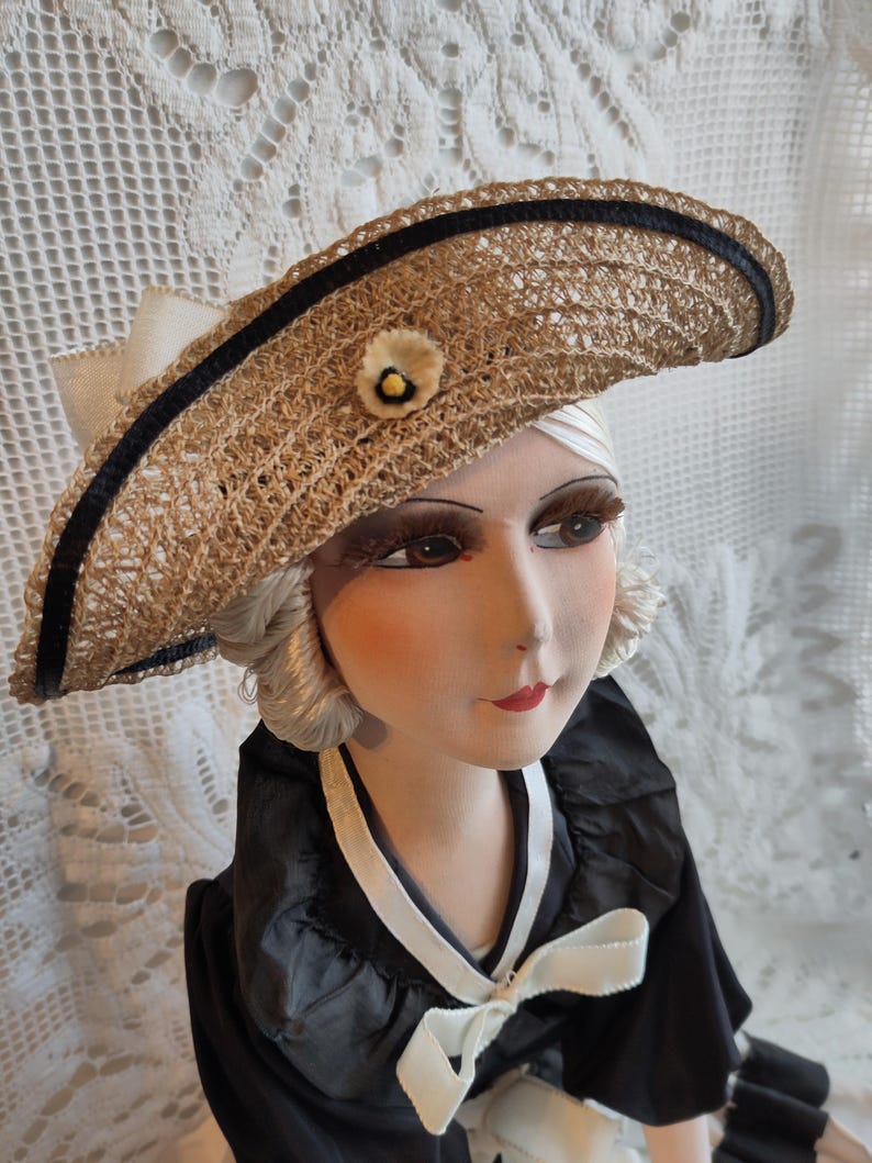 May include: A vintage straw hat with a black ribbon trim and a small floral accent. The hat is part of a doll wearing a black dress with a white bow. The doll has a painted face with red lips and long eyelashes.