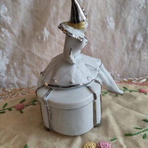 Puede incluir: Una caja de porcelana blanca con ribete plateado. La tapa presenta una figurita de un buf&oacute;n con un sombrero puntiagudo y un traje plateado y amarillo.