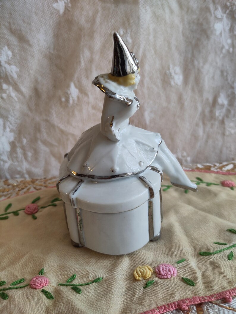 Puede incluir: Una caja de porcelana blanca con ribete plateado. La tapa presenta una figurita de un buf&oacute;n con un sombrero puntiagudo y un traje plateado y amarillo.