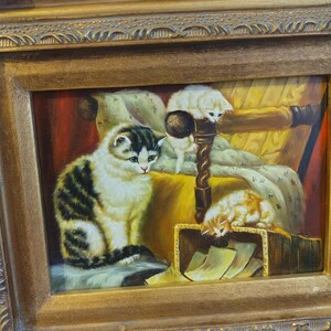 May include: A framed oil painting depicting three cats in a cozy setting. The largest cat is sitting on a patterned cushion, while a smaller white cat is perched on a chair and a third cat is sleeping in a box. The painting is set against a backdrop of a warm, inviting room.