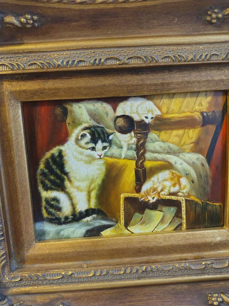 May include: A framed oil painting depicting three cats in a cozy setting. The largest cat is sitting on a patterned cushion, while a smaller white cat is perched on a chair and a third cat is sleeping in a box. The painting is set against a backdrop of a warm, inviting room.