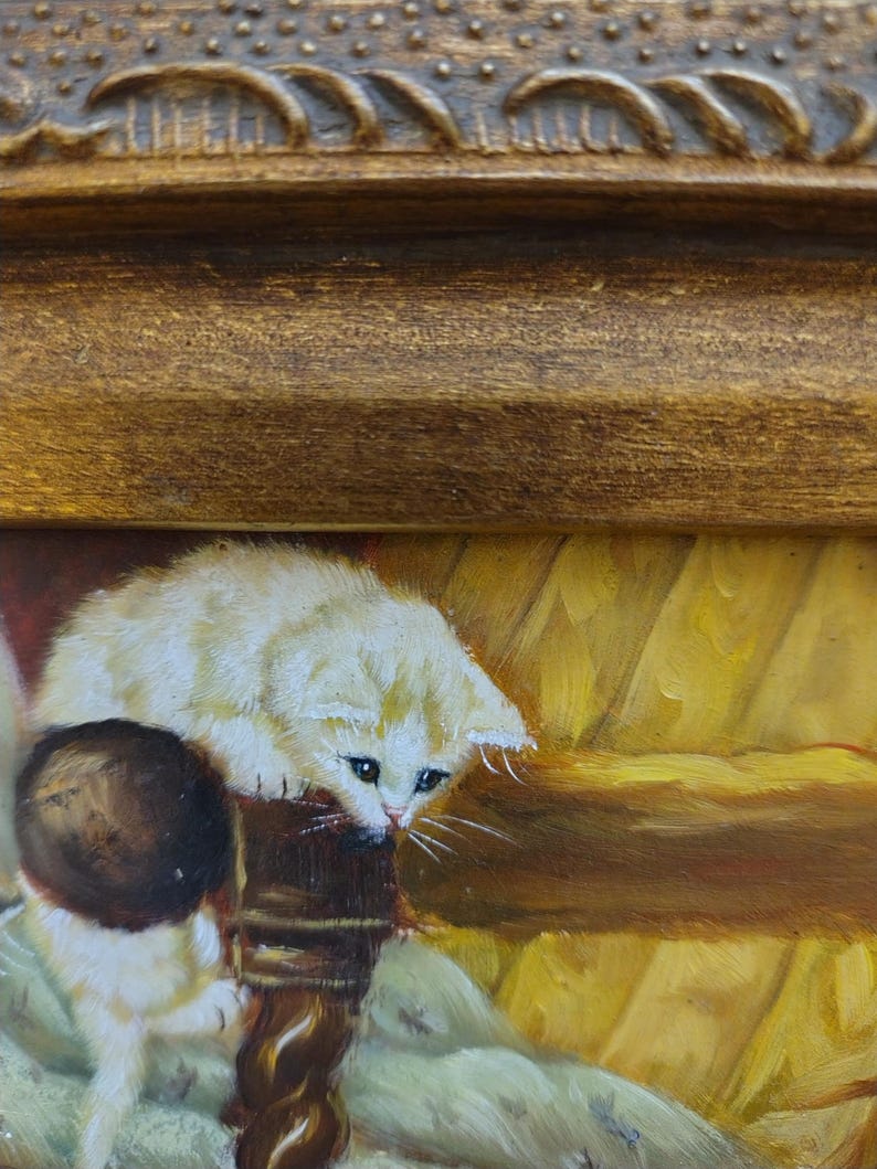 May include: A white kitten with brown markings sits on a wooden chair with a brown ball in its paws. The kitten is looking down at the ball. The background is a yellow and brown painting.