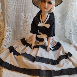 May include: A vintage doll in a black and white dress and a straw hat. The dress features black trim and a white bow. The doll is seated on a white lace surface.