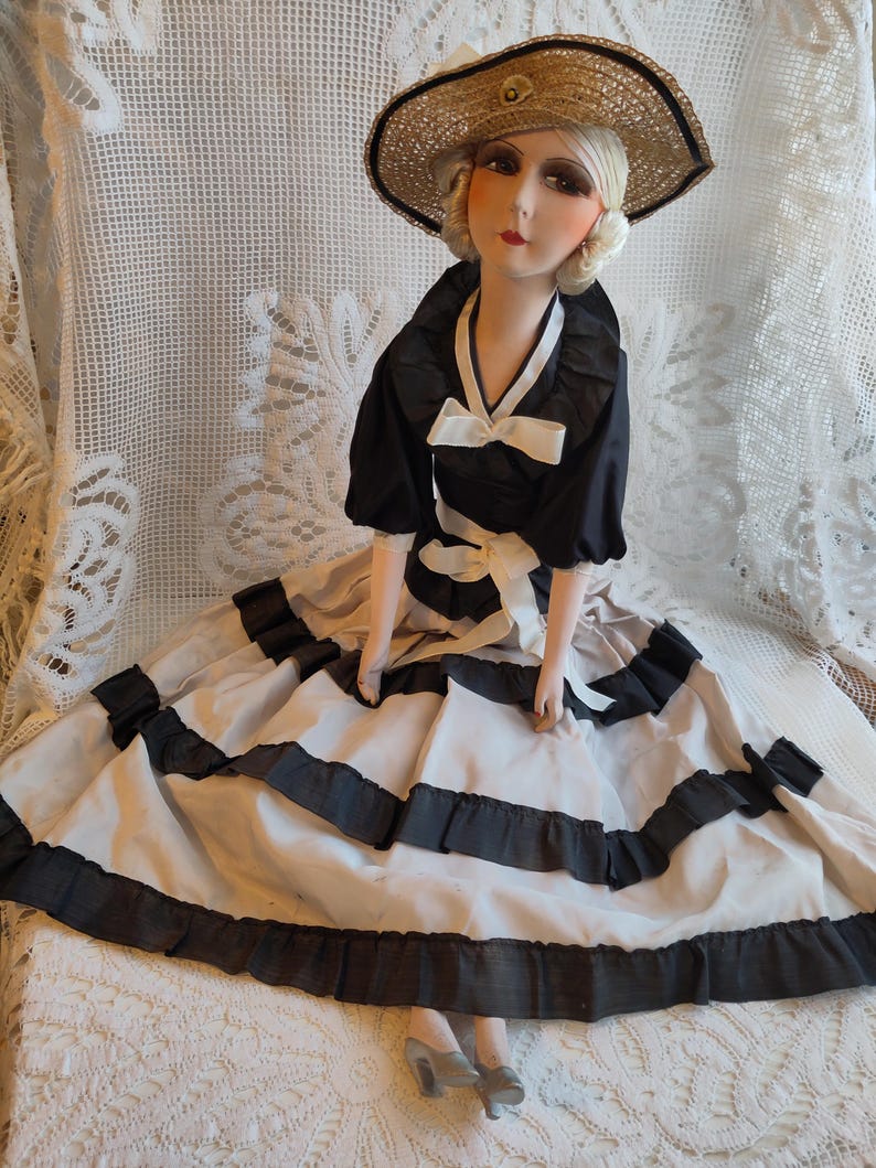 May include: A vintage doll in a black and white dress and a straw hat. The dress features black trim and a white bow. The doll is seated on a white lace surface.