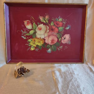 Könnte beinhalten: Ein rechteckiges, weinrotes Serviertablett mit goldenem Rand, verziert mit einem handgemalten Blumenarrangement aus Rosen in Rosa, Weiß und Rot. Neben dem Tablett befindet sich eine Metallrosen-Skulptur mit goldenen Blättern.
