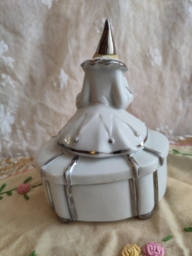 Puede incluir: Caja de porcelana blanca con ribete plateado y tapa con forma de buf&oacute;n. El buf&oacute;n lleva un sombrero puntiagudo plateado con una banda amarilla.