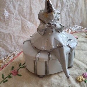 Puede incluir: Una caja de porcelana blanca con ribete plateado. Una figurita de porcelana de un payaso con un sombrero plateado se sienta encima de la caja.