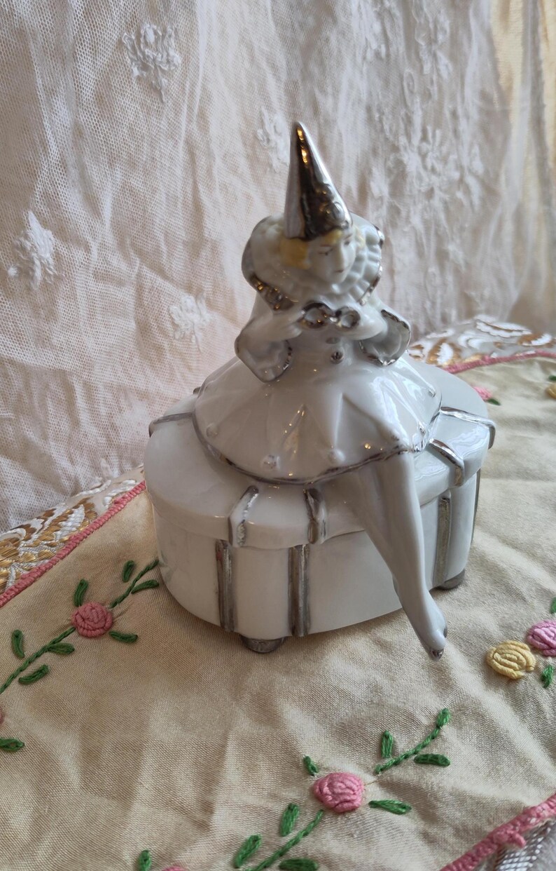 Puede incluir: Una caja de porcelana blanca con ribete plateado. Una figurita de porcelana de un payaso con un sombrero plateado se sienta encima de la caja.