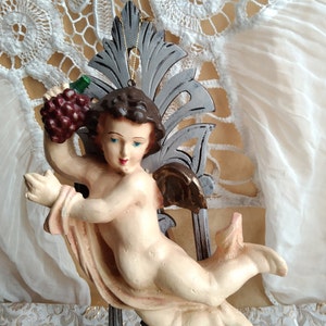 Op de afbeelding: Een cherubijnengelbeeldje dat een tros druiven vasthoudt. De engel is geschilderd in een licht perzikkleurige kleur en heeft bruin haar en blauwe ogen. De engel zit op een grijze en gouden sierlijke basis.