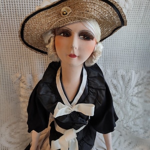 May include: A vintage doll wearing a straw hat with a black band and a small flower. The doll is dressed in a black dress with white bows and accents. The doll's face is painted with detailed features, including long eyelashes and red lips.