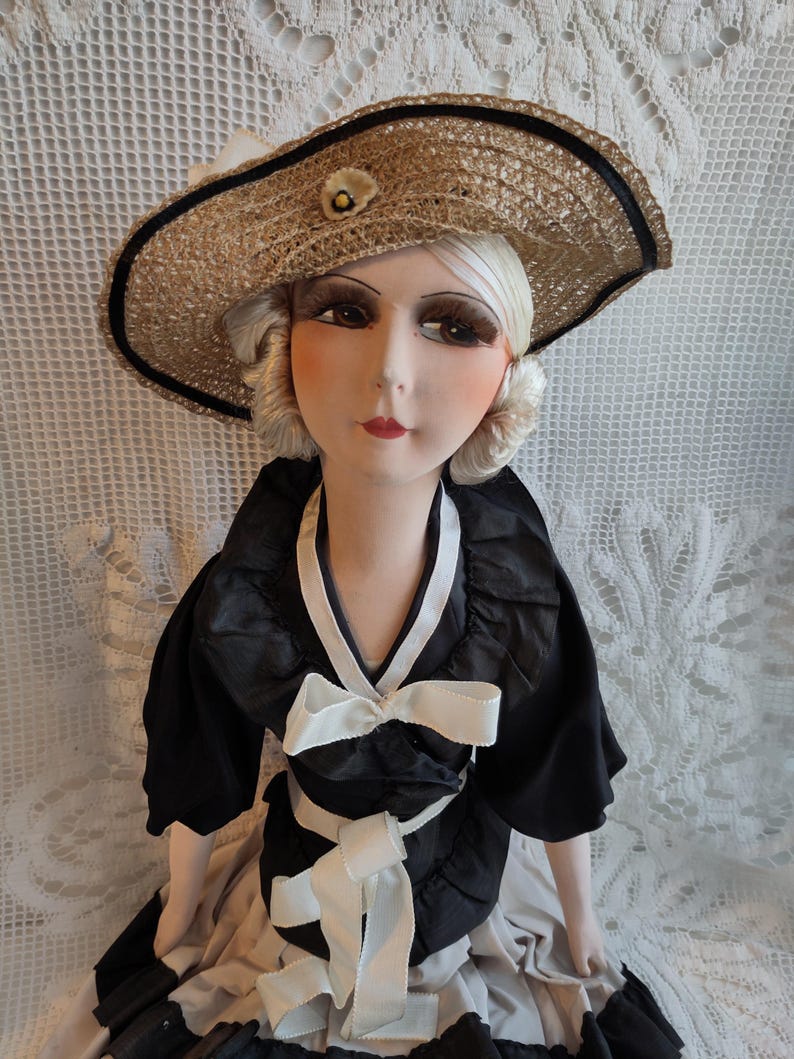 May include: A vintage doll wearing a straw hat with a black band and a small flower. The doll is dressed in a black dress with white bows and accents. The doll's face is painted with detailed features, including long eyelashes and red lips.