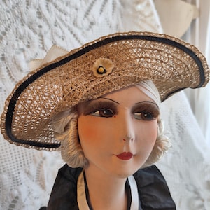 May include: A vintage straw hat with a wide brim, trimmed with a black ribbon and a small flower. The hat is worn by a mannequin head with painted features, including red lips and long eyelashes. The mannequin is wearing a black and white dress.