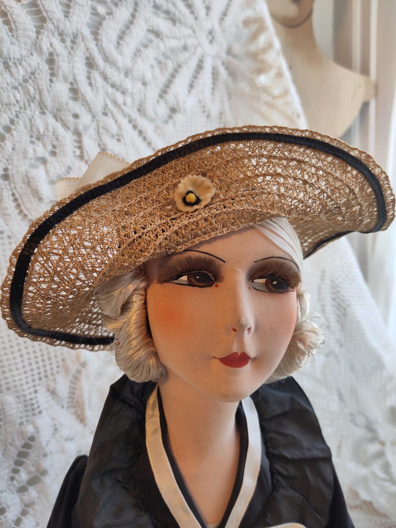May include: A vintage straw hat with a wide brim, trimmed with a black ribbon and a small flower. The hat is worn by a mannequin head with painted features, including red lips and long eyelashes. The mannequin is wearing a black and white dress.