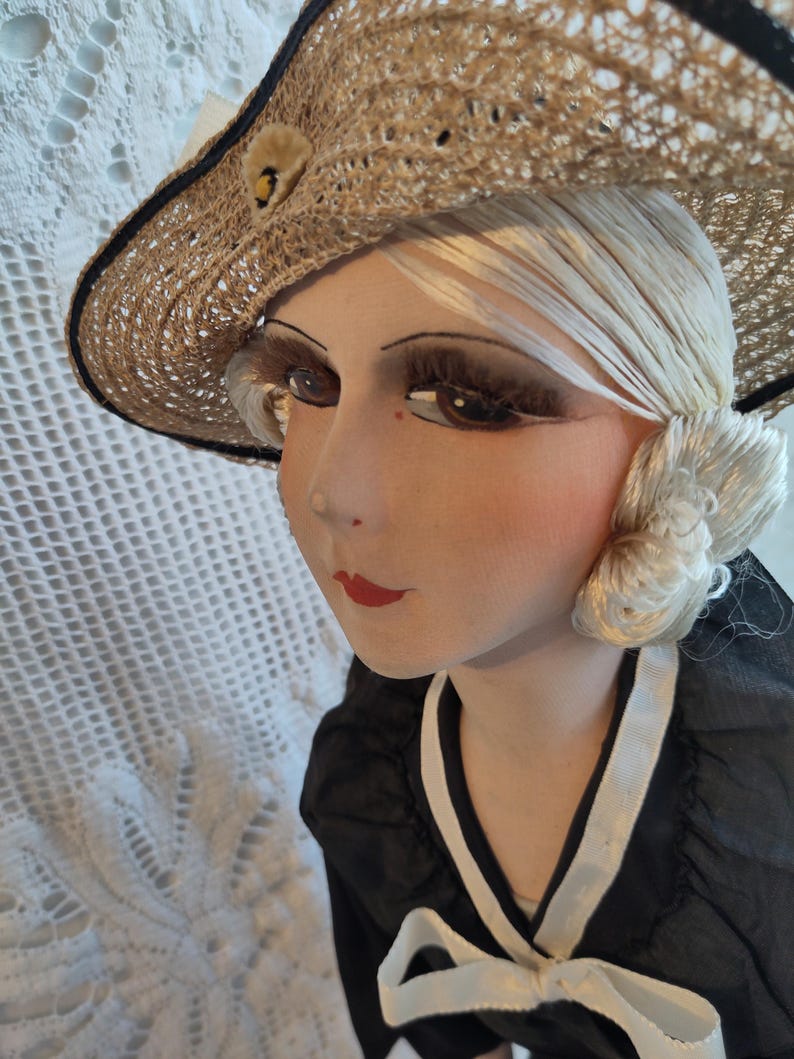 Peut inclure: Poupée vintage avec un chapeau de paille, une robe noire et des cheveux blonds. La poupée a des traits de visage peints, dont des lèvres rouges et de longs cils. Le chapeau est orné d'une bande bleu foncé et d'un petit élément décoratif.