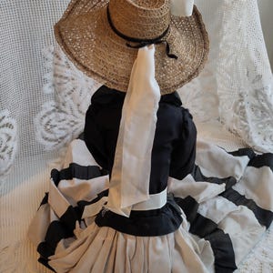 May include: A vintage doll in a straw hat with a black ribbon and a white bow. The doll is wearing a black and white striped dress with a white sash and a black top. The dress has a full skirt.