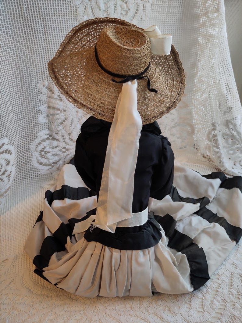May include: A vintage doll in a straw hat with a black ribbon and a white bow. The doll is wearing a black and white striped dress with a white sash and a black top. The dress has a full skirt.