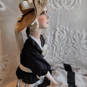 May include: A mannequin head wearing a straw hat with black and navy trim, white ribbon, and a gold-coloured accent. The mannequin is dressed in a black top with a white collar and a long, light-coloured skirt with black ribbon accents.