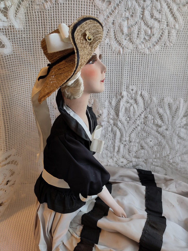 May include: A mannequin head wearing a straw hat with black and navy trim, white ribbon, and a gold-coloured accent. The mannequin is dressed in a black top with a white collar and a long, light-coloured skirt with black ribbon accents.