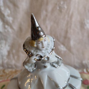 Puede incluir: Una figurita de porcelana blanca de un payaso con un sombrero plateado y ribetes plateados en el traje. El payaso est&aacute; sentado sobre una base de porcelana blanca.
