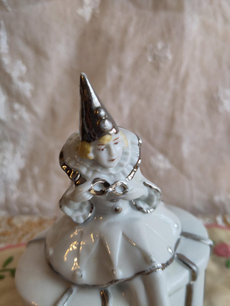 Puede incluir: Una figurita de porcelana blanca de un payaso con un sombrero plateado y ribetes plateados en el traje. El payaso est&aacute; sentado sobre una base de porcelana blanca.