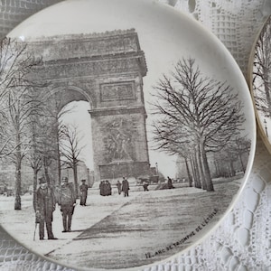 May include: Two white ceramic plates with a black and white illustration of the Arc de Triomphe in Paris. The plates feature the text "12. Arc de Triomphe de l'Etoile" along the rim, and depict figures and trees.