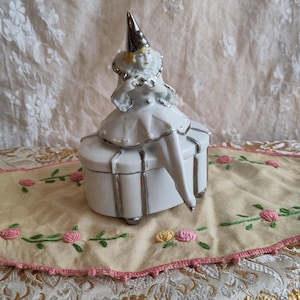 Puede incluir: Una caja de porcelana blanca con ribete plateado. Una figurita de porcelana de una mujer con un sombrero de buf&oacute;n plateado y blanco se sienta encima de la caja.