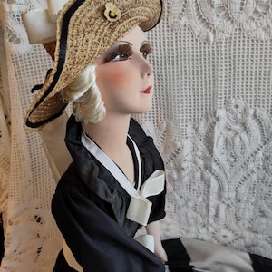May include: A vintage doll wearing a straw hat with a black trim and a small flower. The doll is dressed in a black dress with white trim and a white bow. The doll's face is painted with rosy cheeks and red lips.