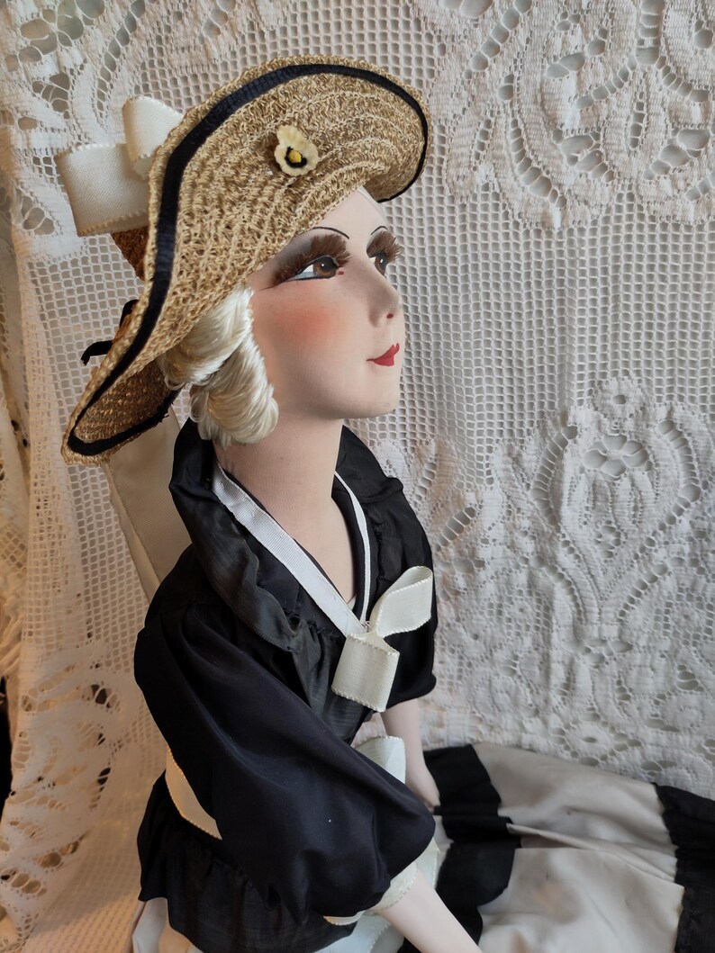 May include: A vintage doll wearing a straw hat with a black trim and a small flower. The doll is dressed in a black dress with white trim and a white bow. The doll's face is painted with rosy cheeks and red lips.