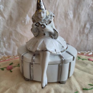 Puede incluir: Una caja de porcelana blanca con ribete plateado. Una figurita de porcelana de una mujer con un sombrero de buf&oacute;n y sosteniendo un par de gafas se sienta encima de la caja.
