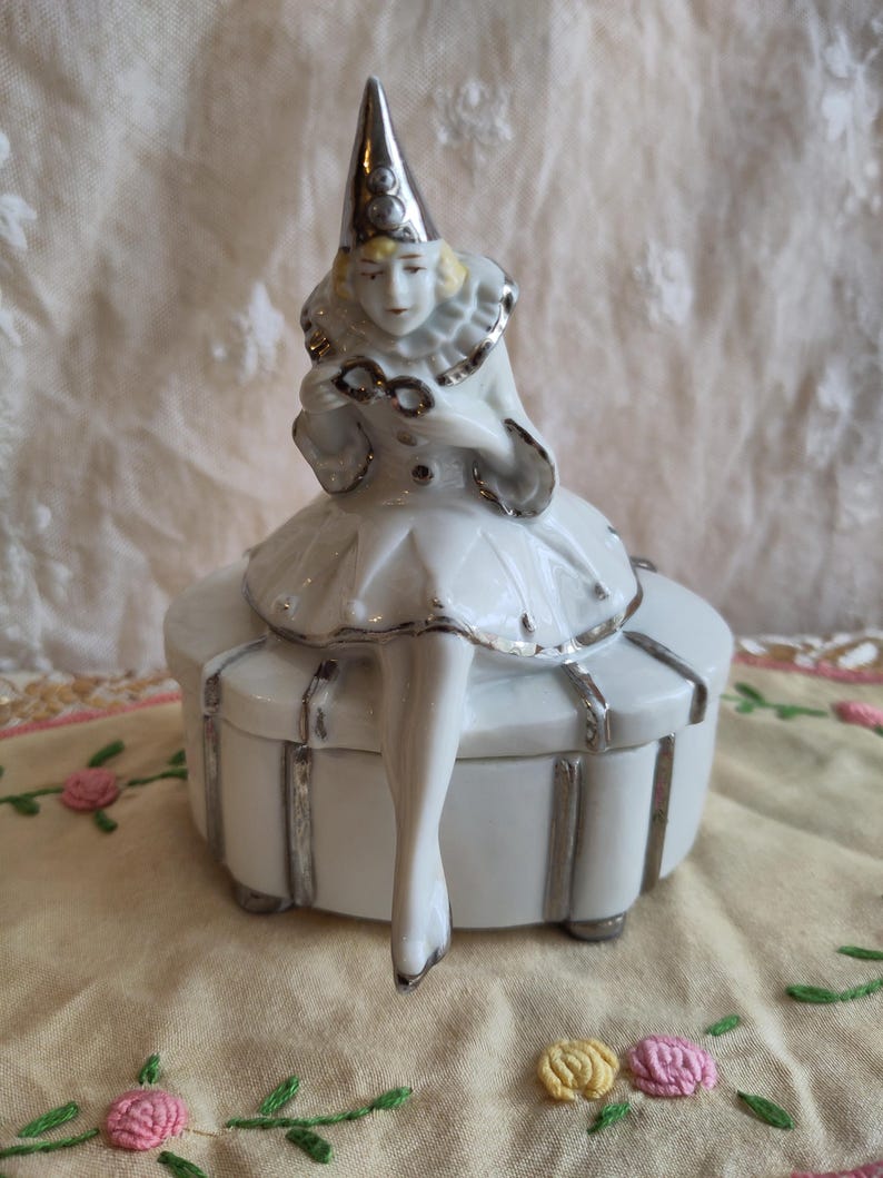 Puede incluir: Una caja de porcelana blanca con ribete plateado. Una figurita de porcelana de una mujer con un sombrero de buf&oacute;n y sosteniendo un par de gafas se sienta encima de la caja.