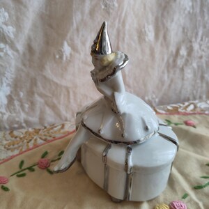 Puede incluir: Una caja de porcelana blanca con ribete plateado. Una figurita de porcelana de una ni&ntilde;a con un sombrero plateado se sienta encima de la caja.