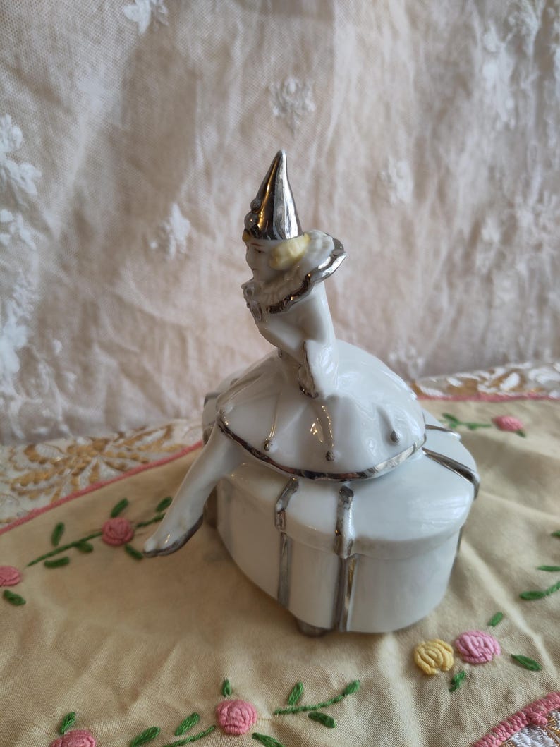 Puede incluir: Una caja de porcelana blanca con ribete plateado. Una figurita de porcelana de una ni&ntilde;a con un sombrero plateado se sienta encima de la caja.