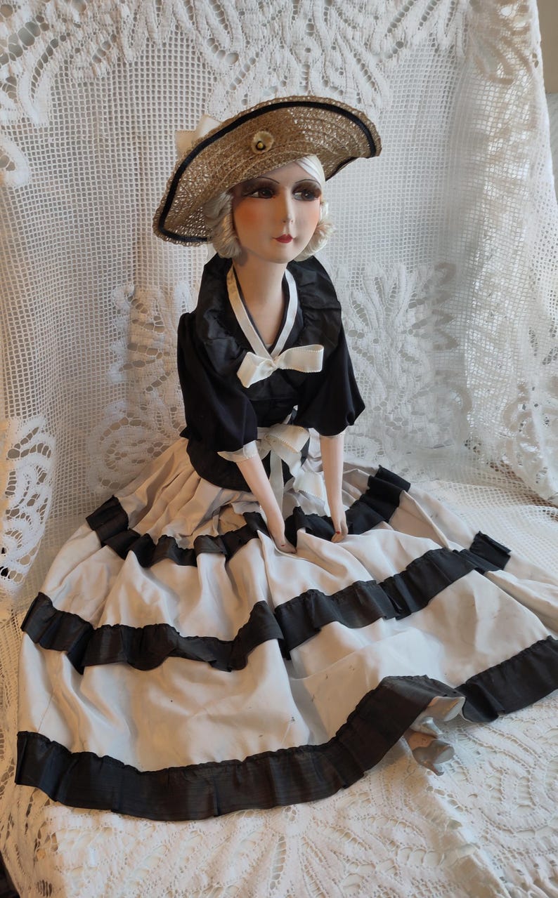 May include: A vintage doll in a black and white dress and a straw hat. The doll's dress features a black bodice and a full skirt with black trim. The hat is straw-coloured with a black band and a decorative embellishment.